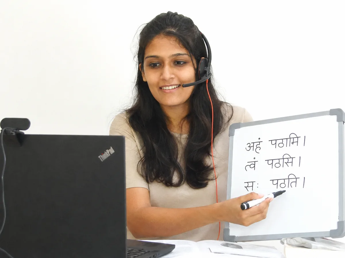 Ms. Ishita Parikh teaching a live online Sanskrit class, with Sanskrit verb forms written on the whiteboard behind her